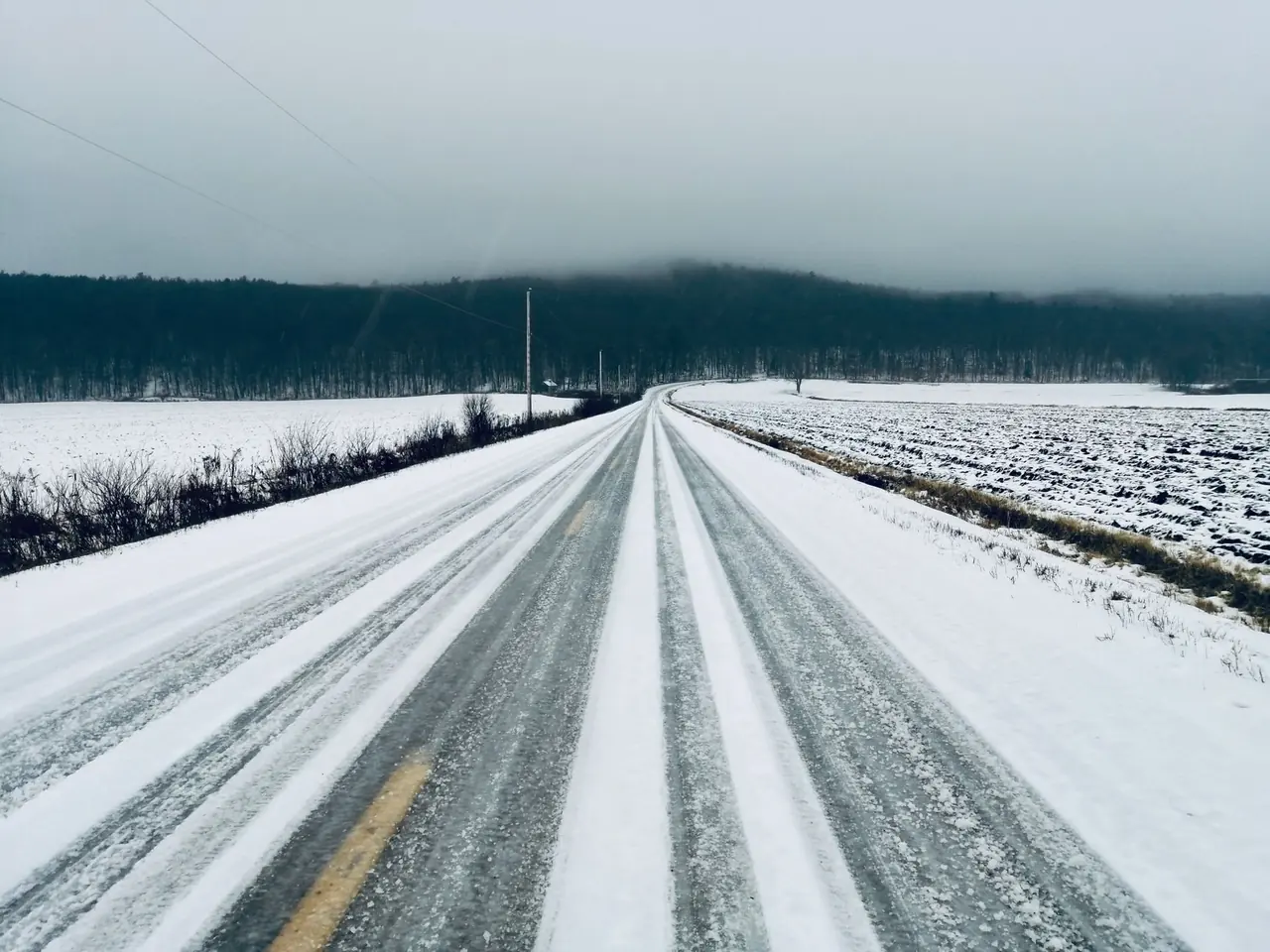Route enneigée rectiligne bordée de champs et de forêt sous un ciel gris
