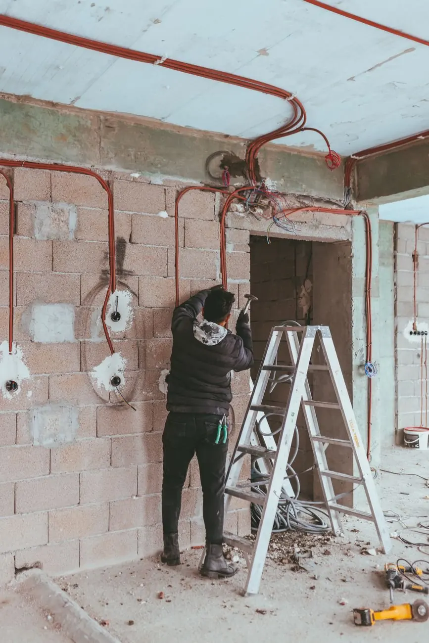 Électricien de dos sur un escabeau installant des conduits rouges dans un local en rénovation